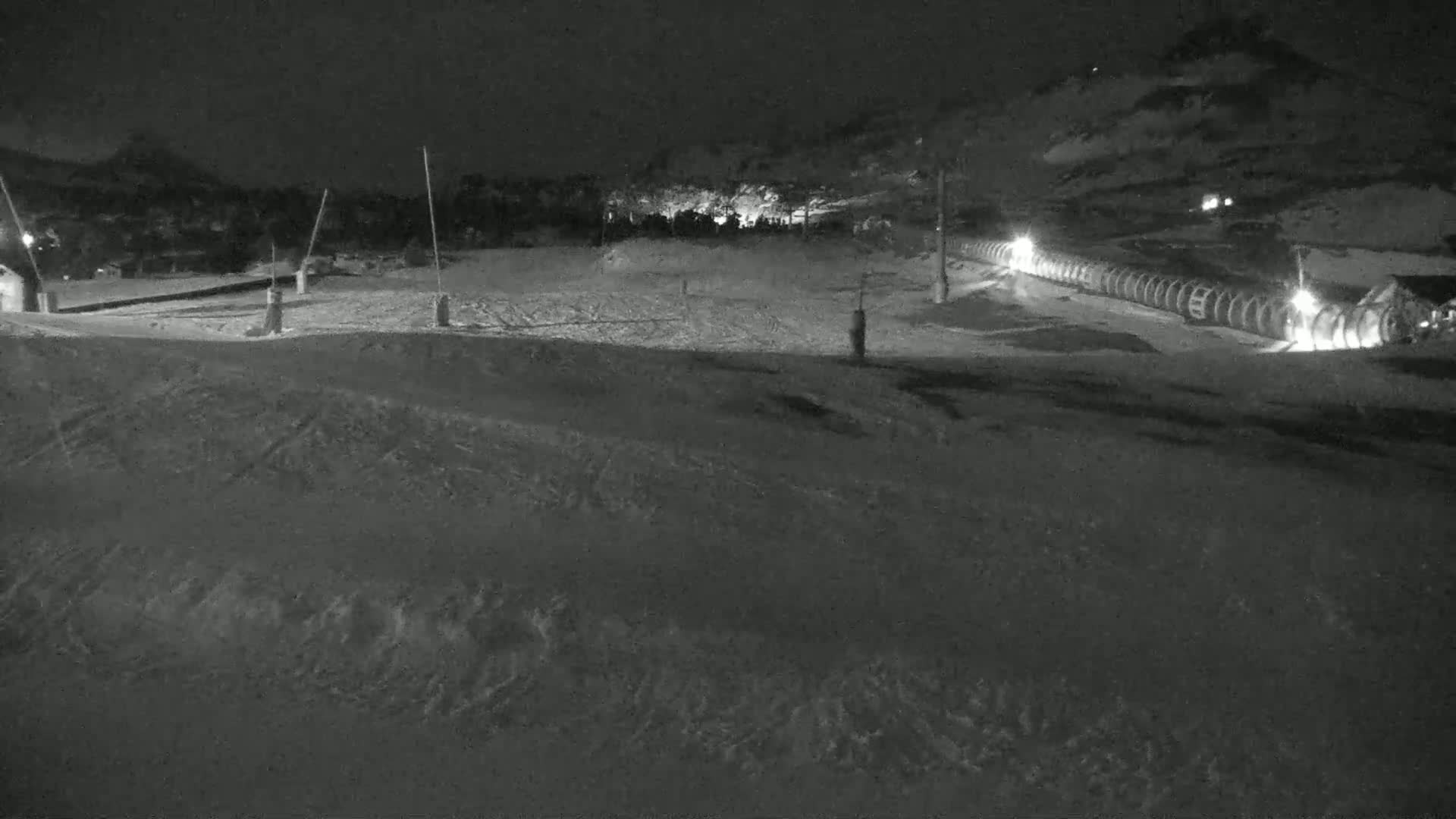 A snow-covered landscape at night, showing a ski lift and some buildings in the distance under dark skies.