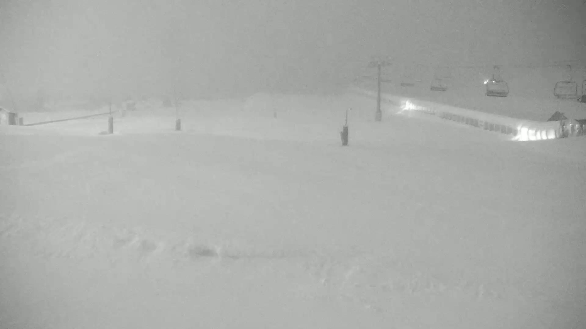 A snow-covered ski resort is seen in heavy snowfall, with a chairlift and several ski lift vehicles visible in the distance.