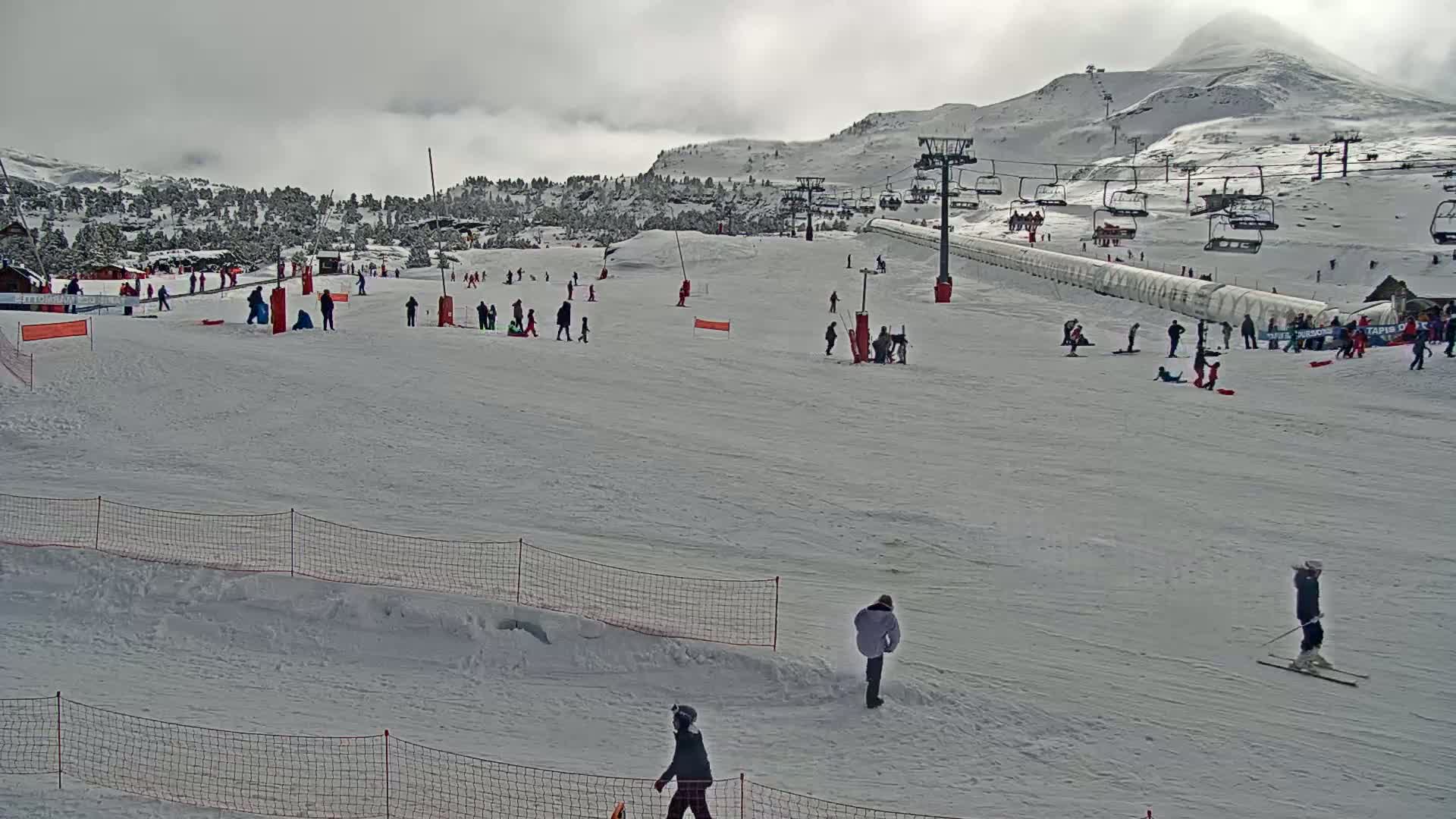 A snowy ski slope with several people skiing, snowboarding, and sledding under an overcast sky.