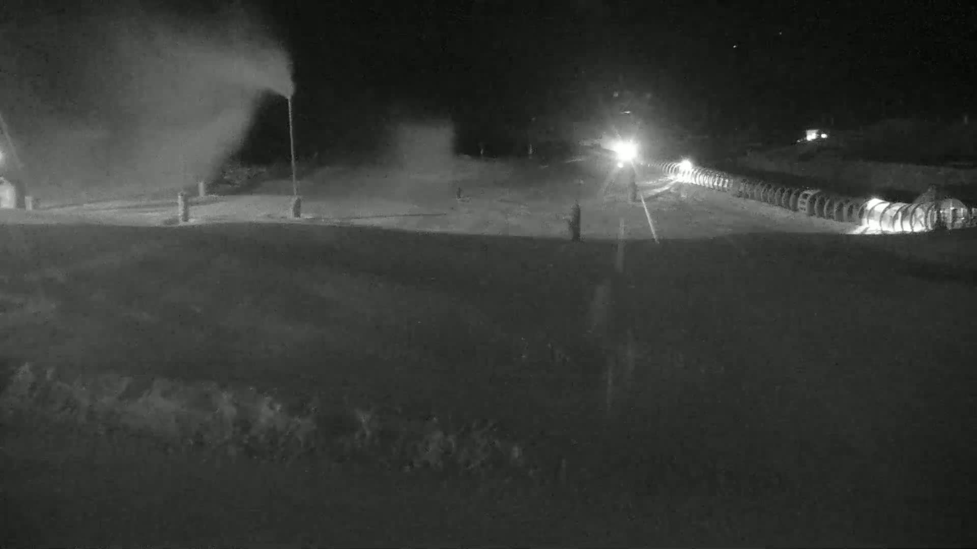 A snowmaking operation is underway at night on a ski slope, with snow guns emitting plumes of artificial snow.