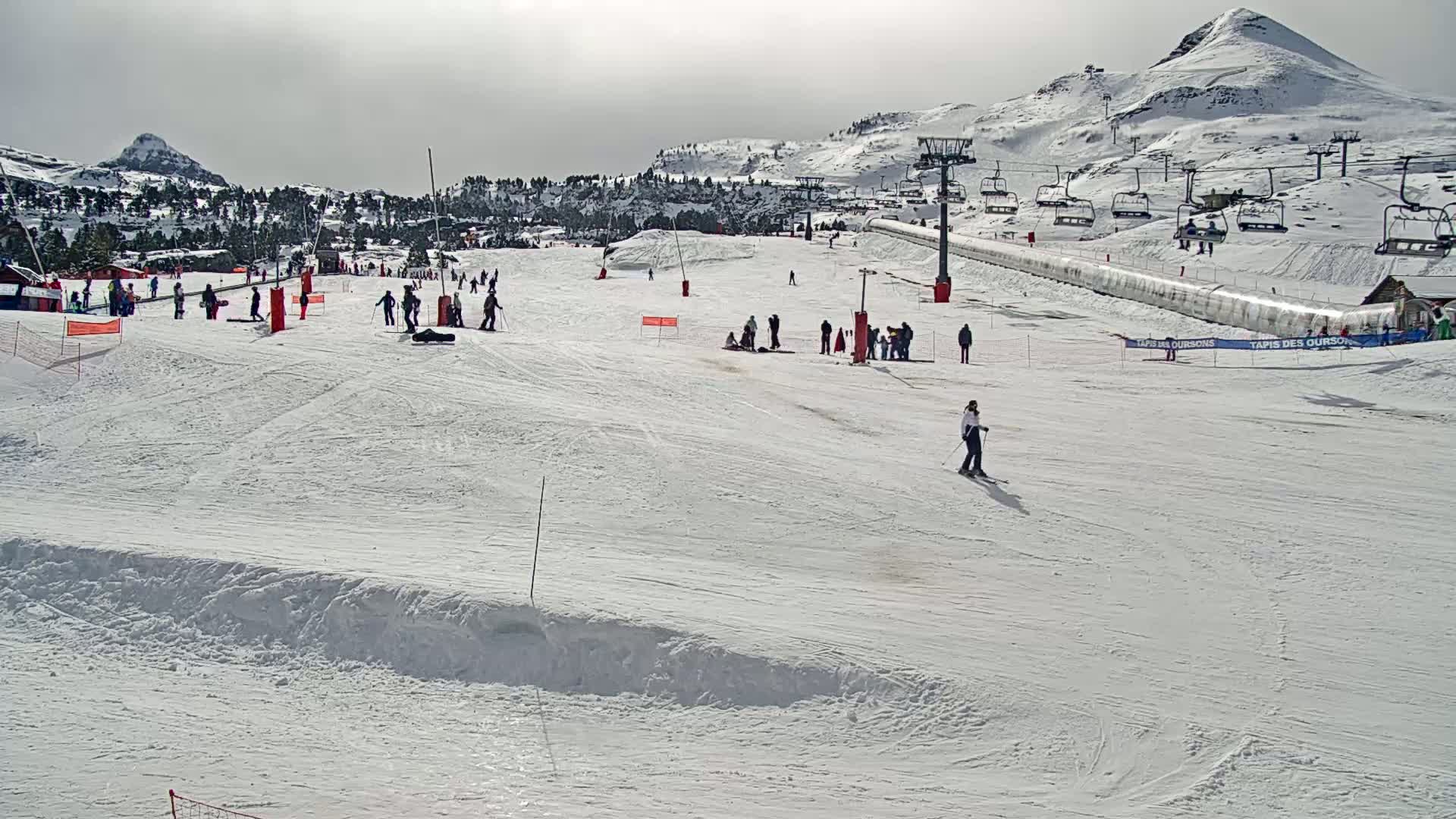 A snow-covered ski slope with several skiers and snowboarders, ski lifts, and a mountain in the background under a mostly sunny sky.