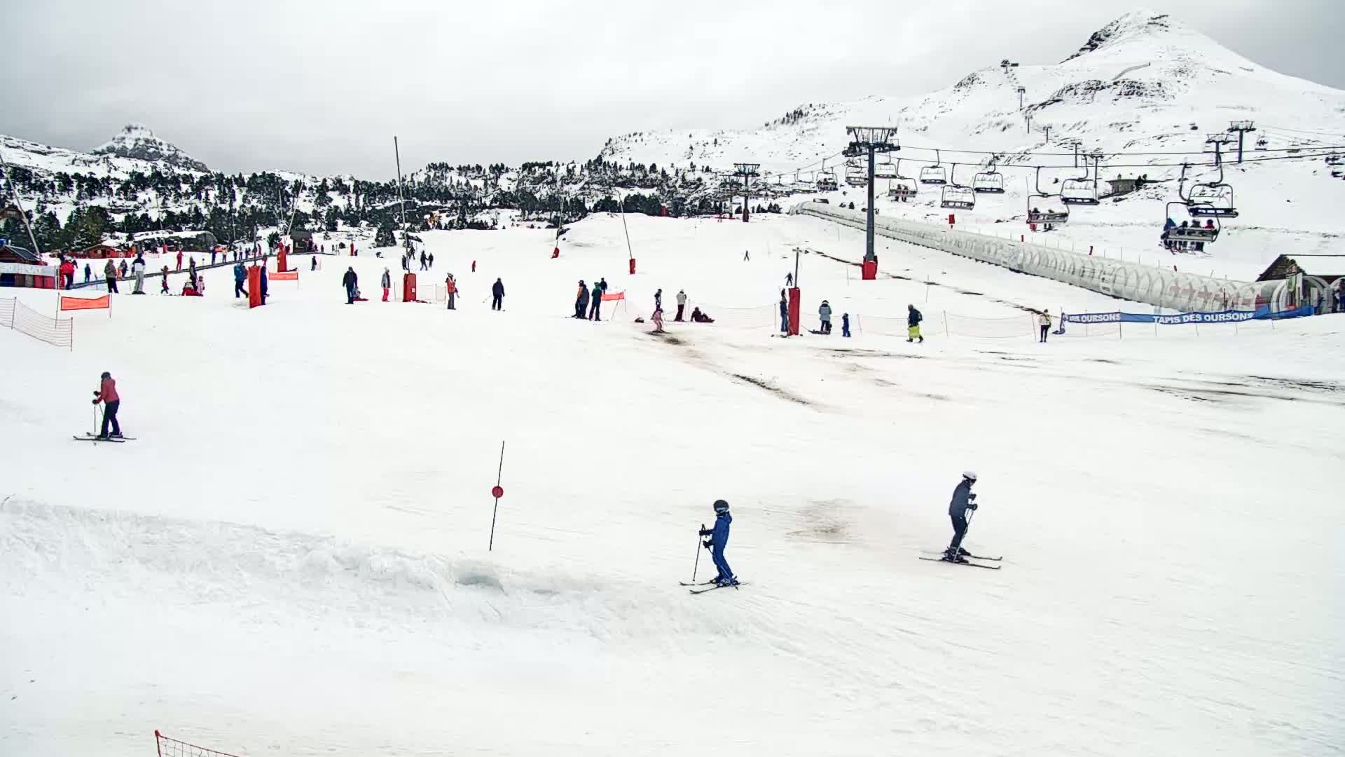 A snowy ski slope with several skiers and snowboarders, chairlifts, and a mountain in the background under overcast skies.