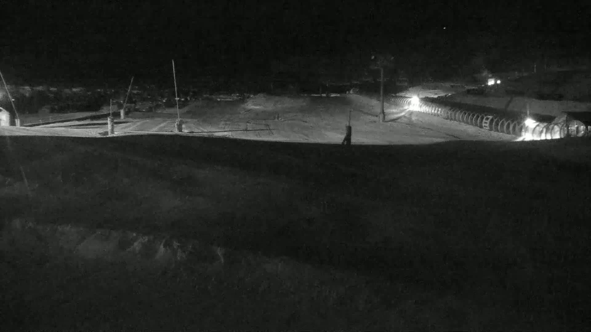 A snow-covered area at night, illuminated by lights, shows a row of objects, possibly snowmaking machines, and other structures in the distance.