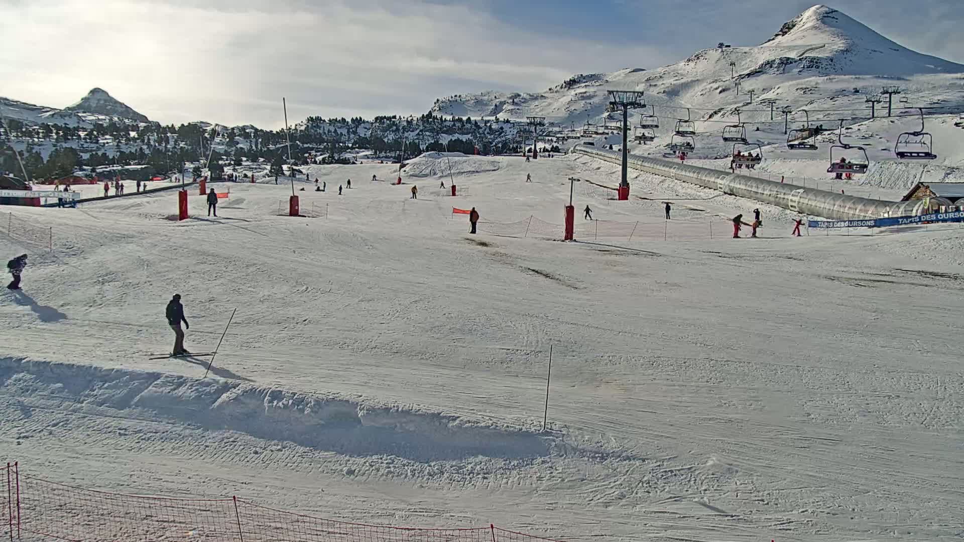 A snowy ski slope with several skiers and snowboarders, a chairlift, and a mountain in the background under sunny conditions.