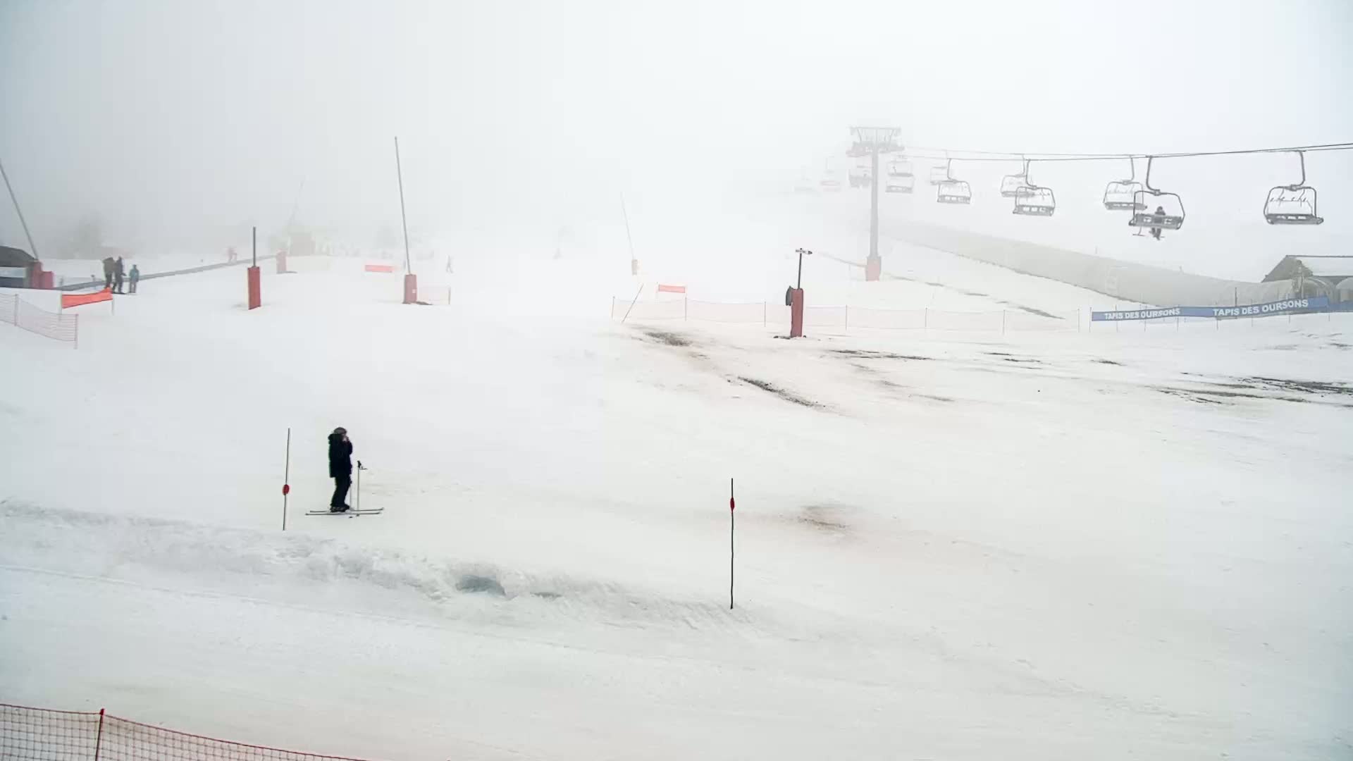 A snowy ski slope is shrouded in fog, with a chairlift visible in the background and a lone skier standing on the slope.