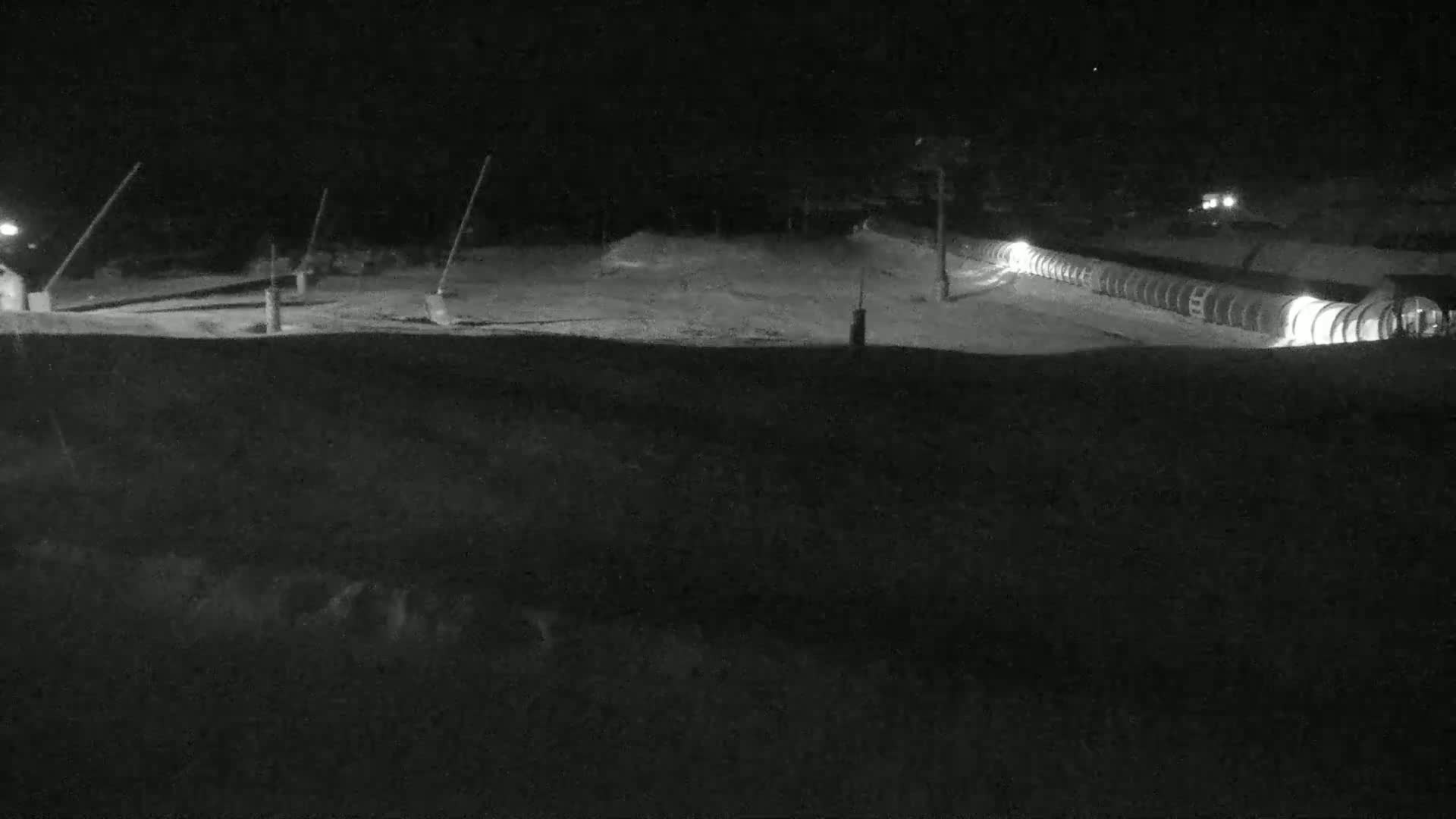 A nighttime, snow-covered scene shows a long row of illuminated structures, possibly snowmaking equipment, situated on a hill.