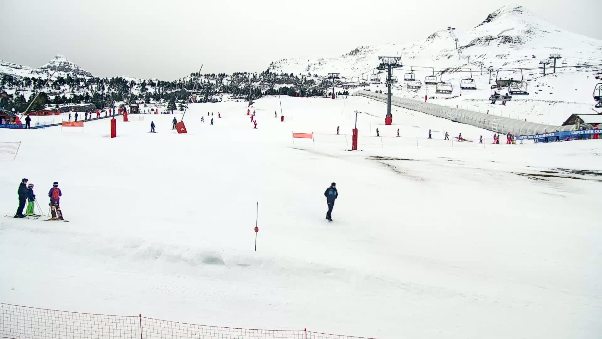 A snowy ski slope with several skiers and snowboarders, chairlifts, and a mountain in the background under an overcast sky.
