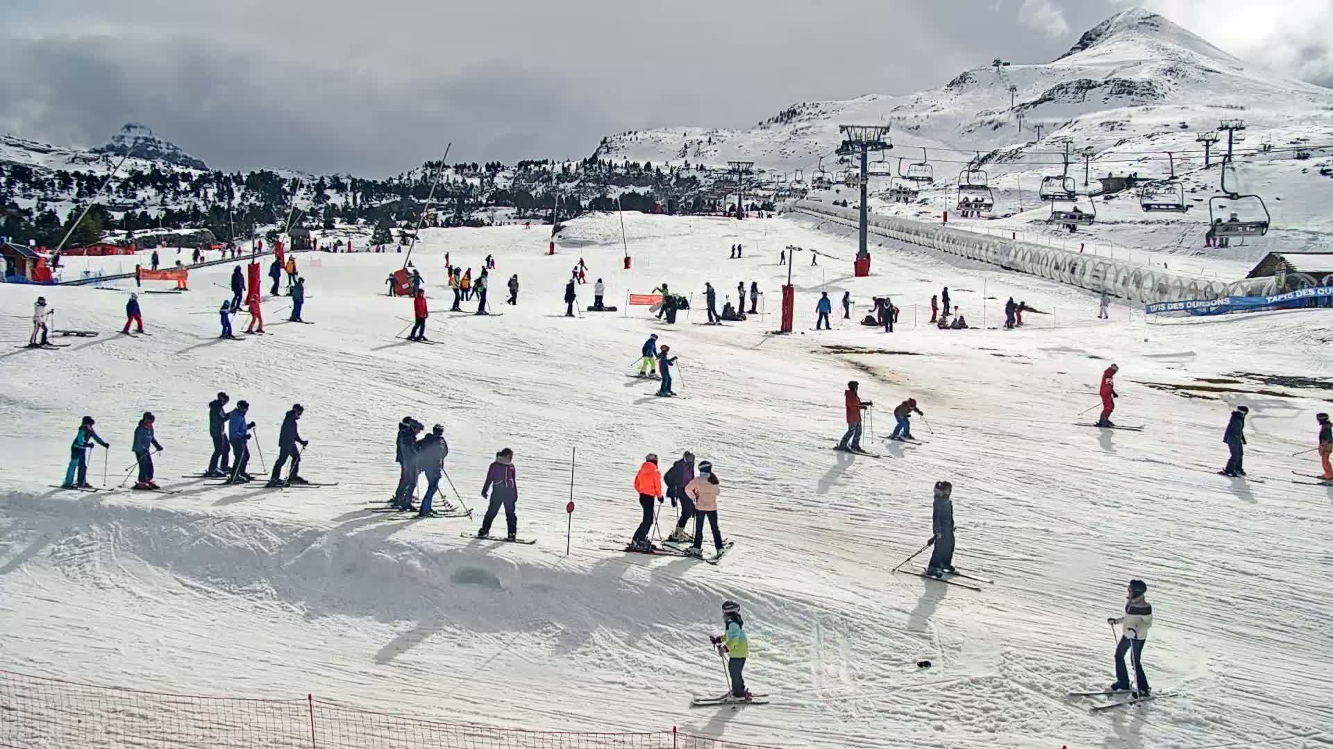 Many skiers and snowboarders are enjoying a snowy day on a ski slope with a mountain in the background under a cloudy sky.
