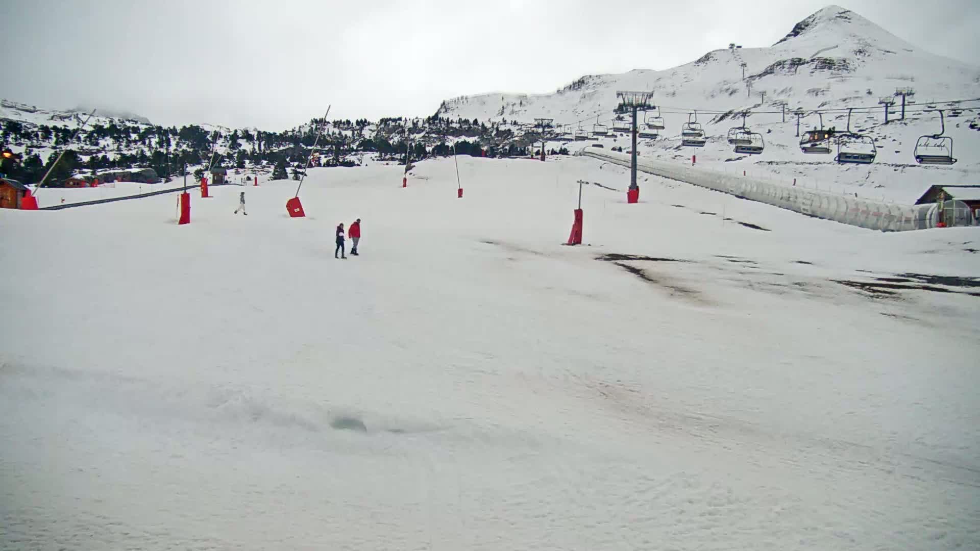 A snow-covered ski slope with a chairlift and several people standing on the piste under an overcast sky.