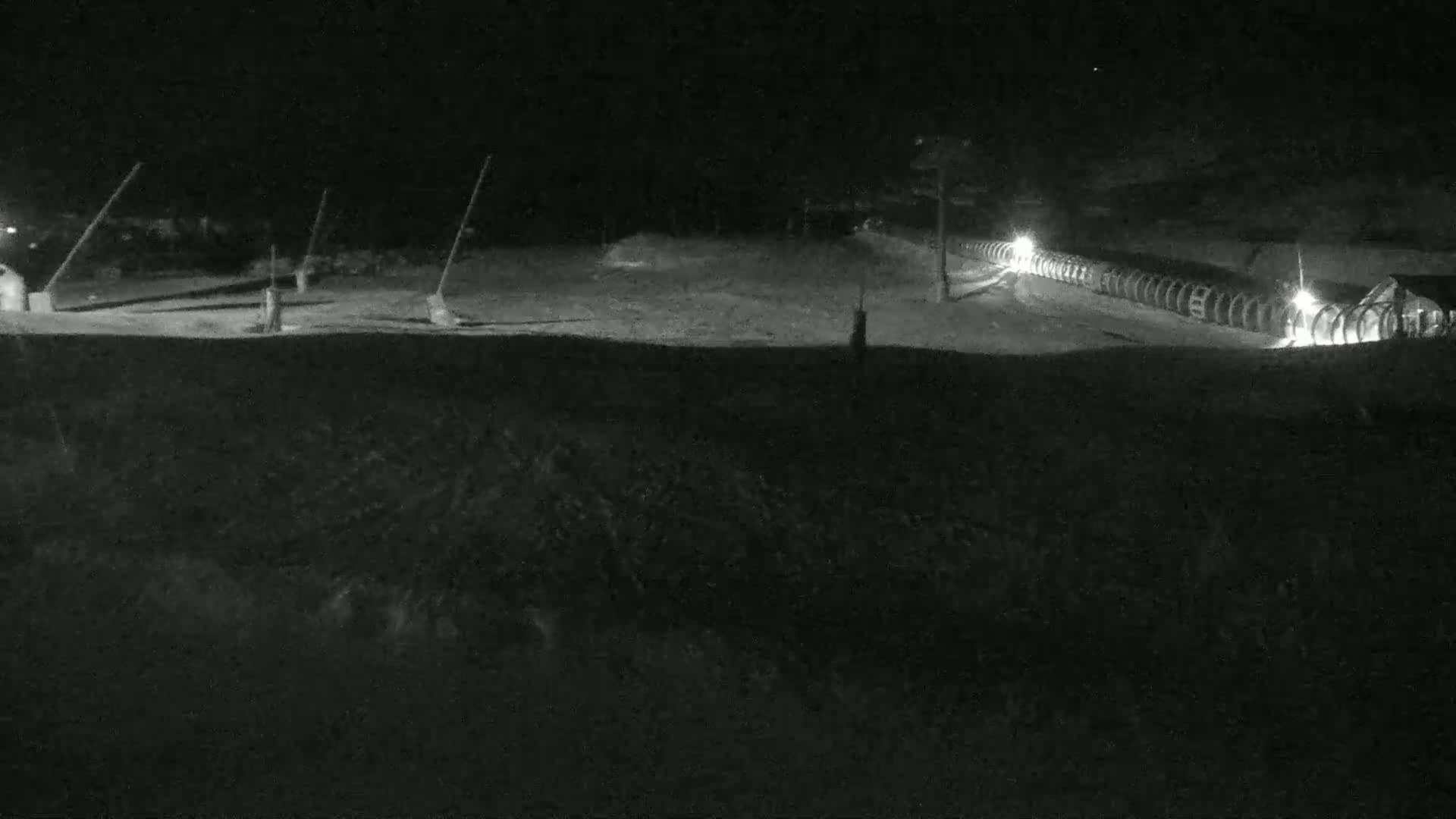 A nighttime, snow-covered scene shows a long line of illuminated objects, possibly snowmaking equipment, extending from a building in the distance.