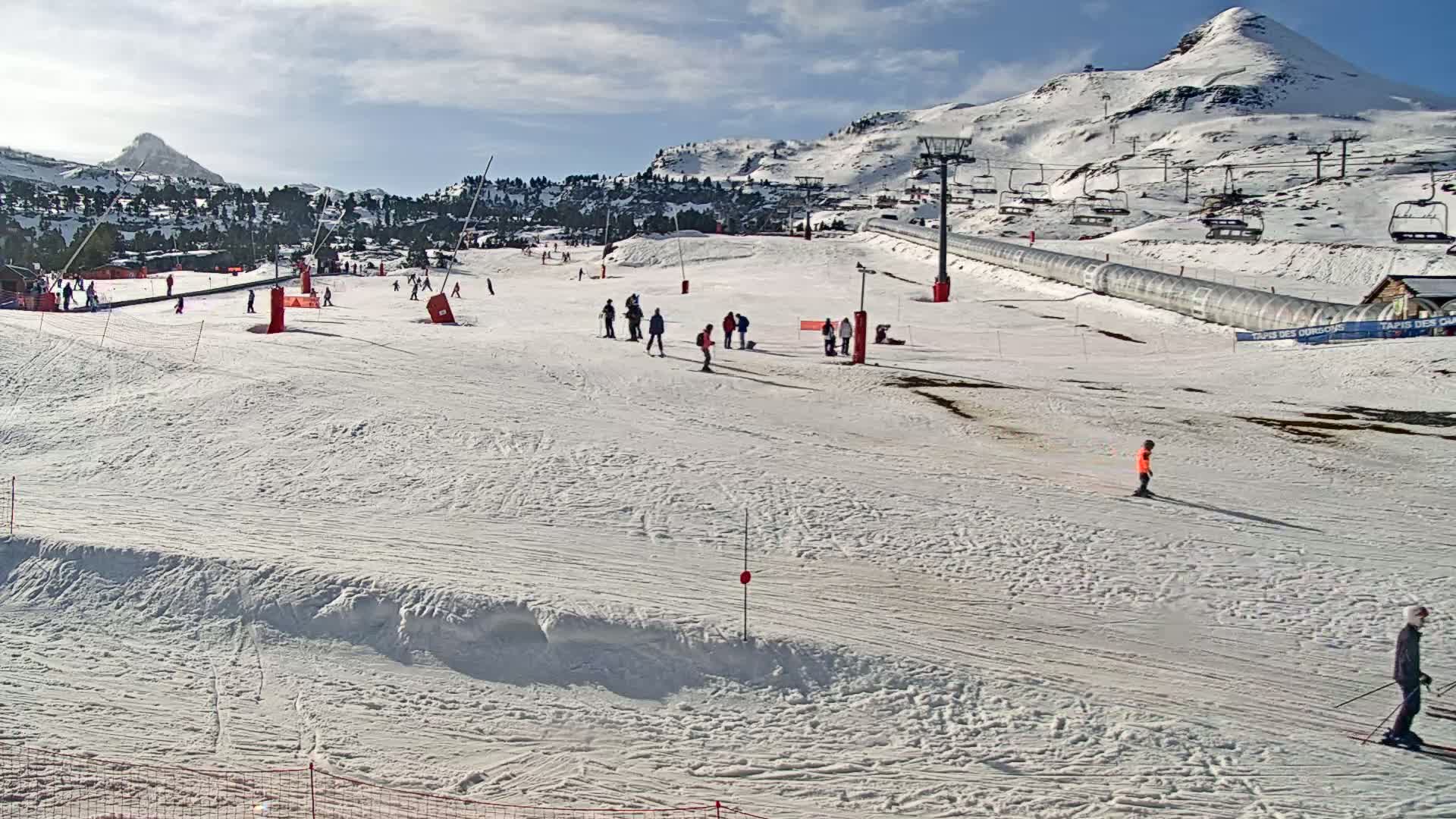 A snow-covered ski slope with several skiers and snowboarders, under a partly cloudy sky, is situated in a mountainous region.