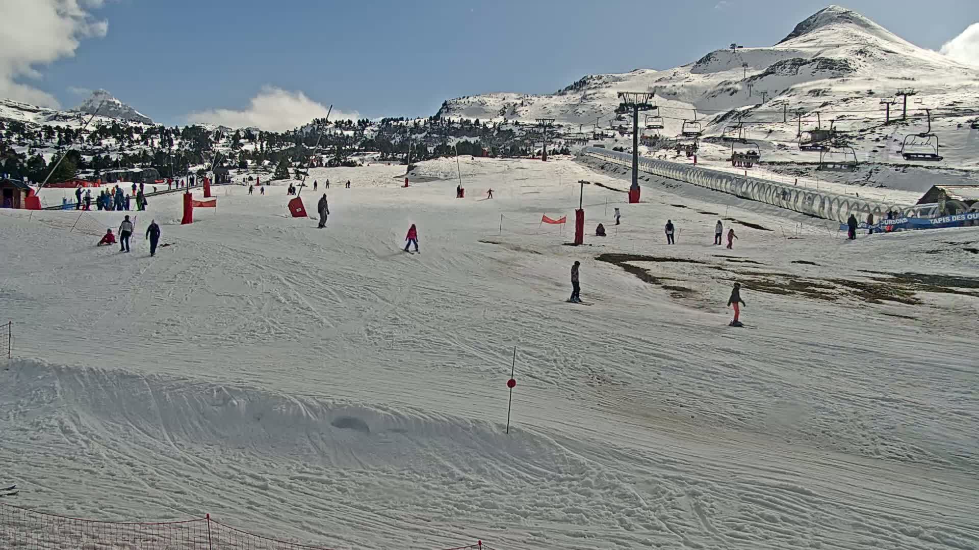 A snow-covered ski slope with several skiers and snowboarders, ski lifts, and a mountain in the background under a mostly sunny sky.