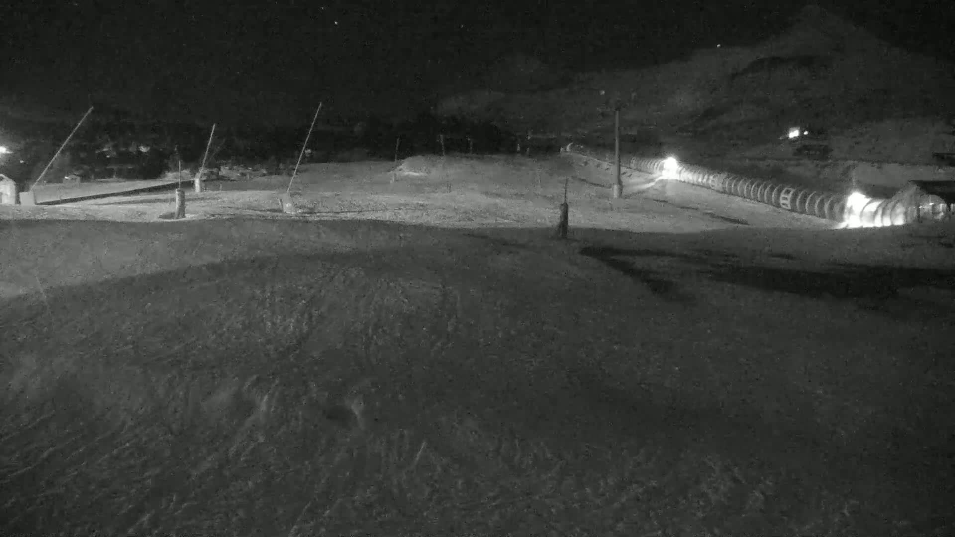 A snow-covered ski slope is illuminated at night by artificial lights, with a ski lift visible in the background.