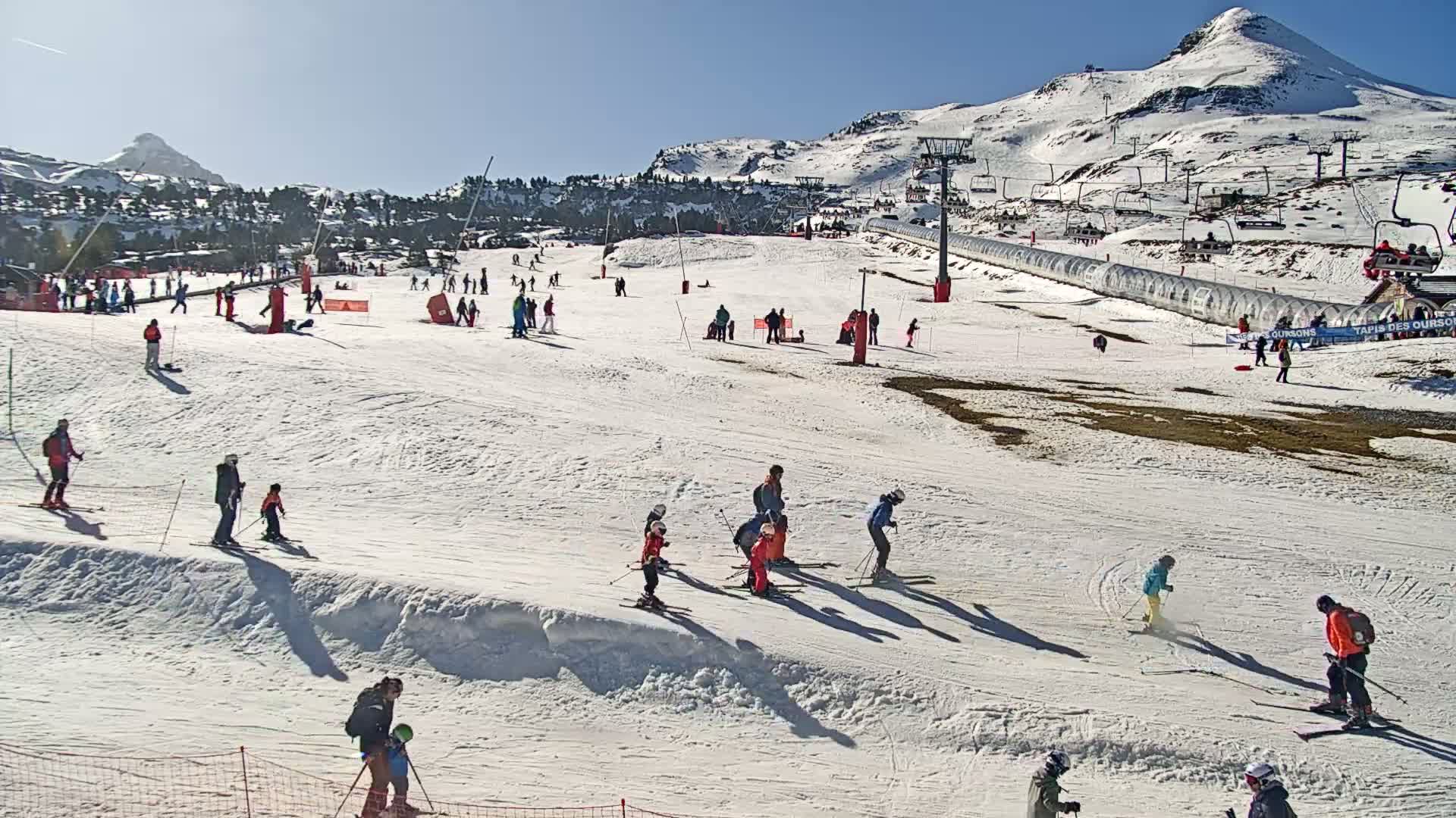 A sunny ski slope with many skiers and snowboarders, some using ski lifts, set against a snow-covered mountain backdrop.