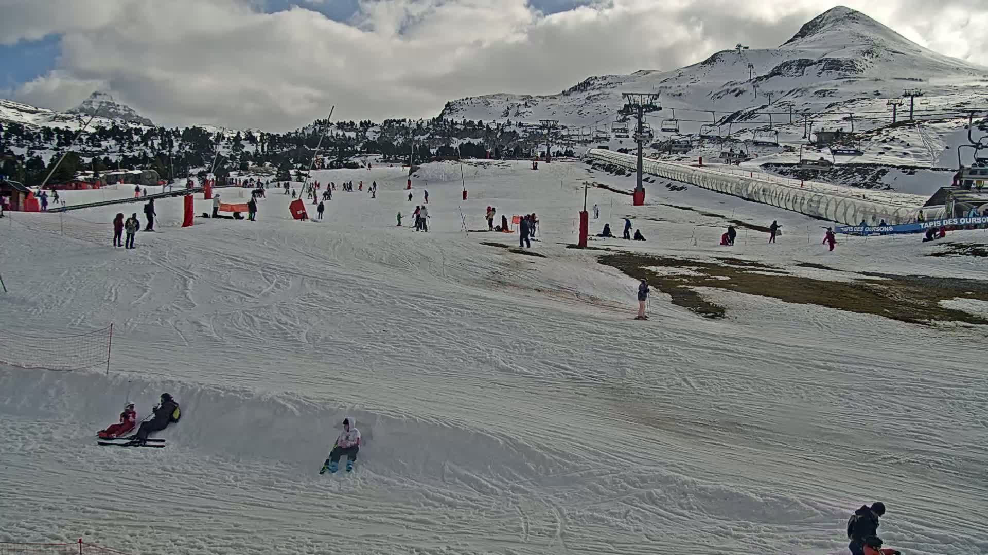 A snow-covered ski slope with scattered skiers and snowboarders under a partly cloudy sky, features a mountain in the background.