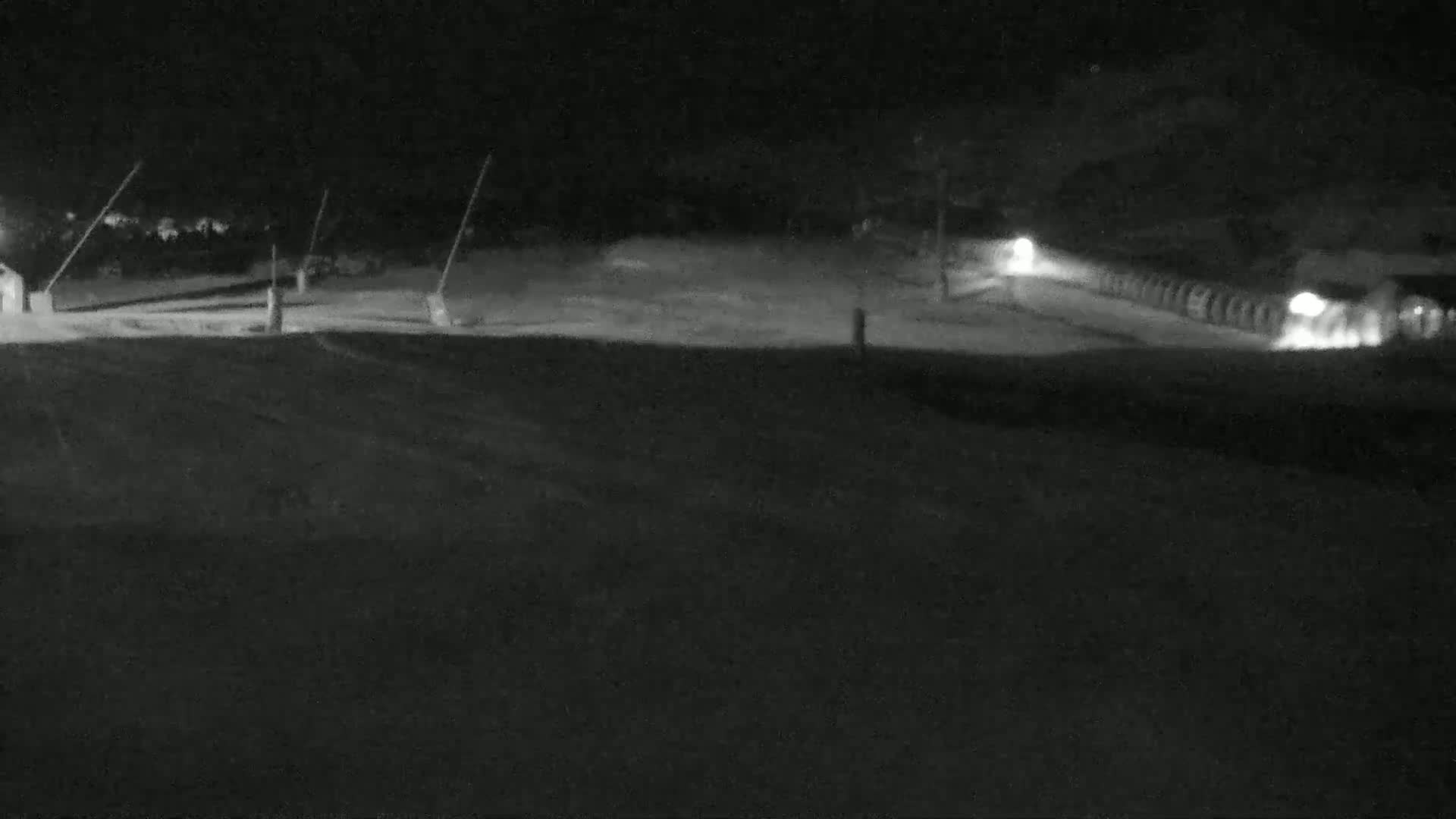 A nighttime, snow-covered scene shows a mostly empty expanse with a few indistinct structures and faint lights in the distance.