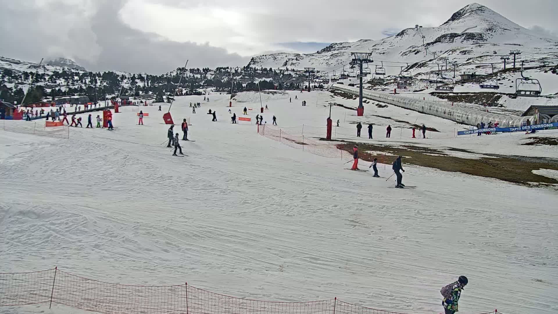 A ski slope with numerous skiers and snowboarders under an overcast sky, situated against a backdrop of snow-covered mountains.