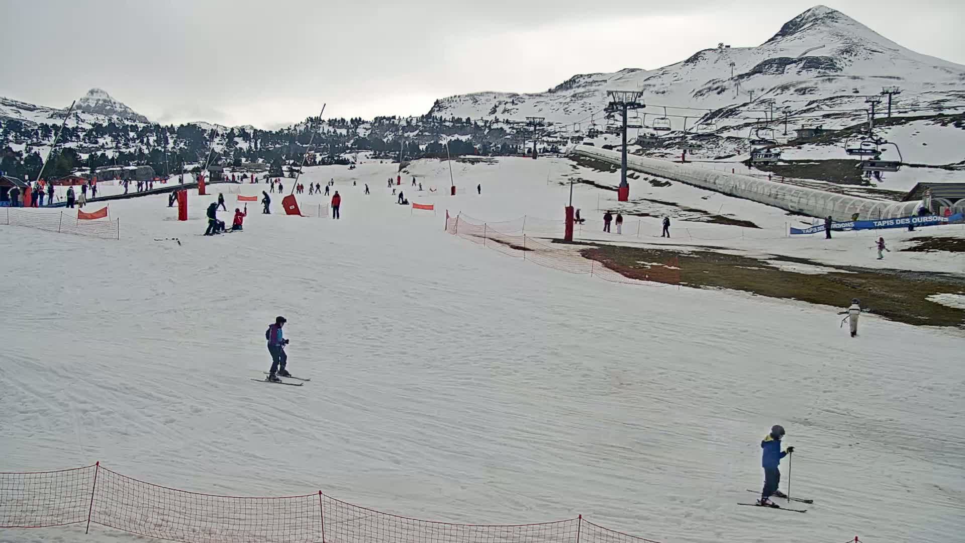 A snowy ski slope with several skiers and snowboarders, ski lifts, and snow-covered mountains in the background under an overcast sky.
