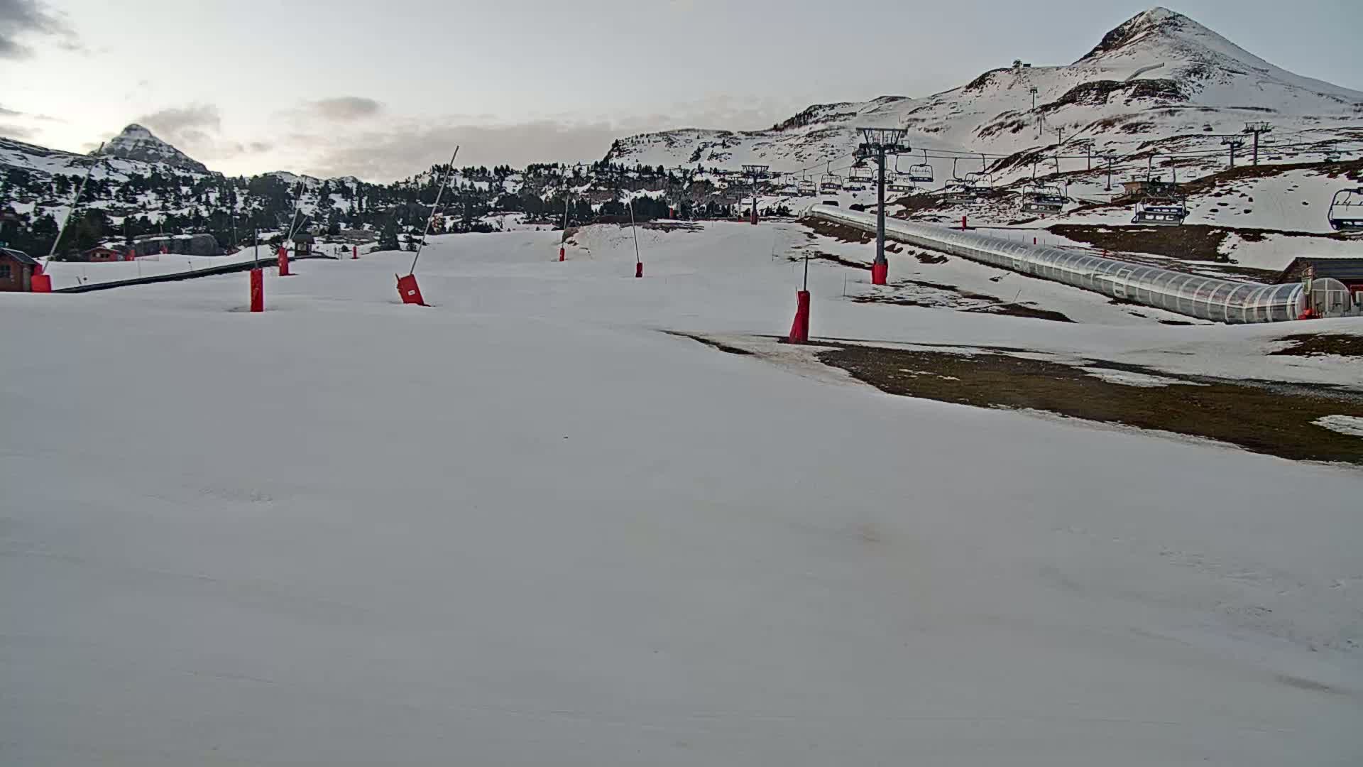 A mostly snow-covered ski slope with a chairlift and covered walkway is situated in front of snow-capped mountains under a cloudy sky.