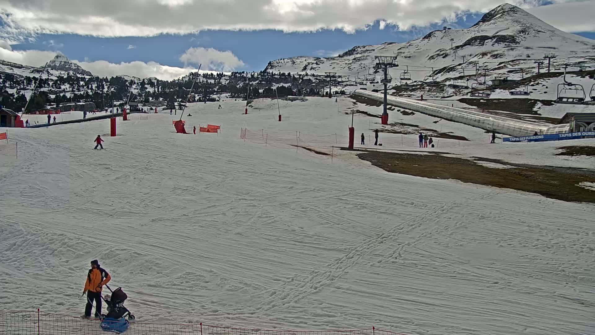 A mostly cloudy day shows a snow-covered ski slope with several skiers and snowboarders, ski lifts, and a person pushing a stroller.