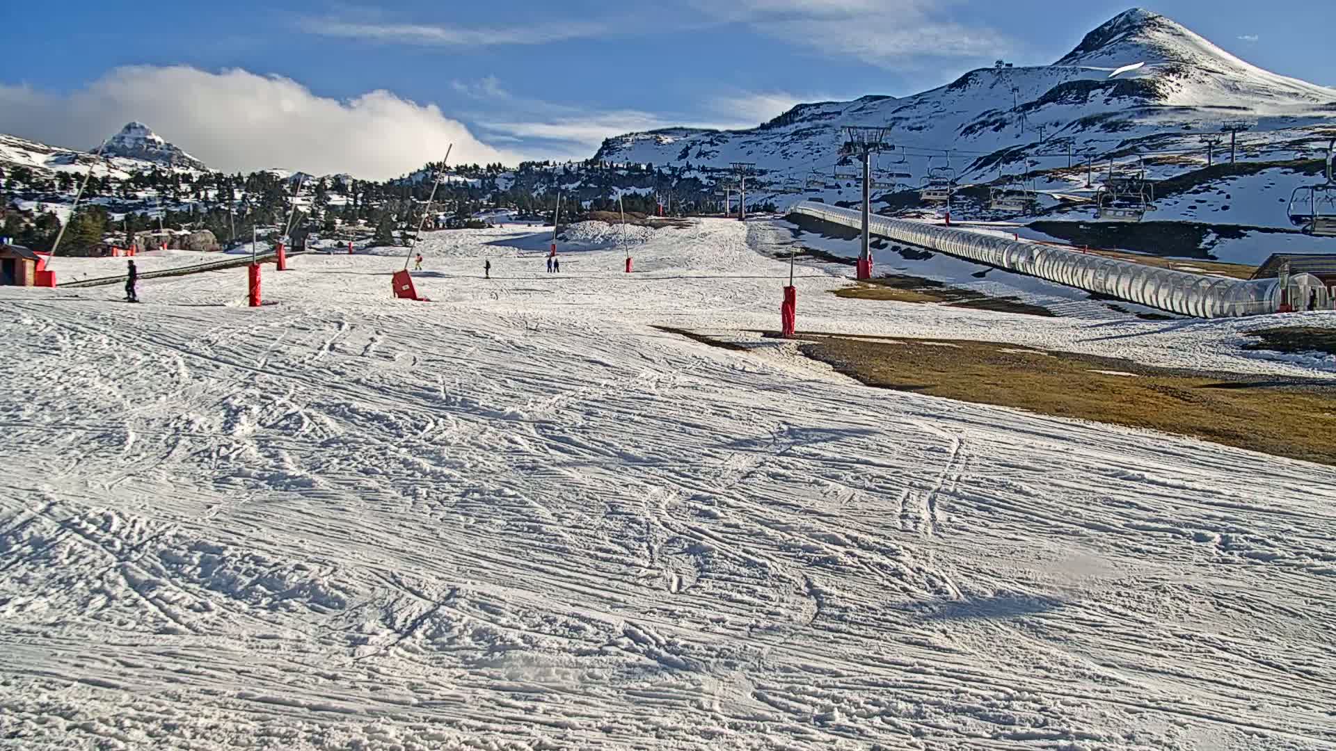 A snow-covered ski slope with several skiers visible, under a partly cloudy blue sky, is nestled among snow-dusted mountains.