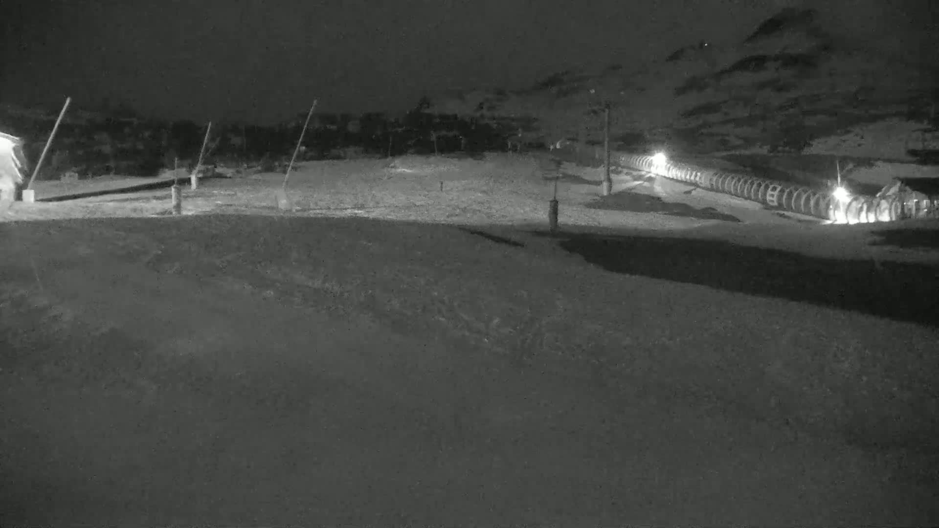 A nighttime, snow-covered ski slope is illuminated by lights along a lift, with a dark hillside in the background.
