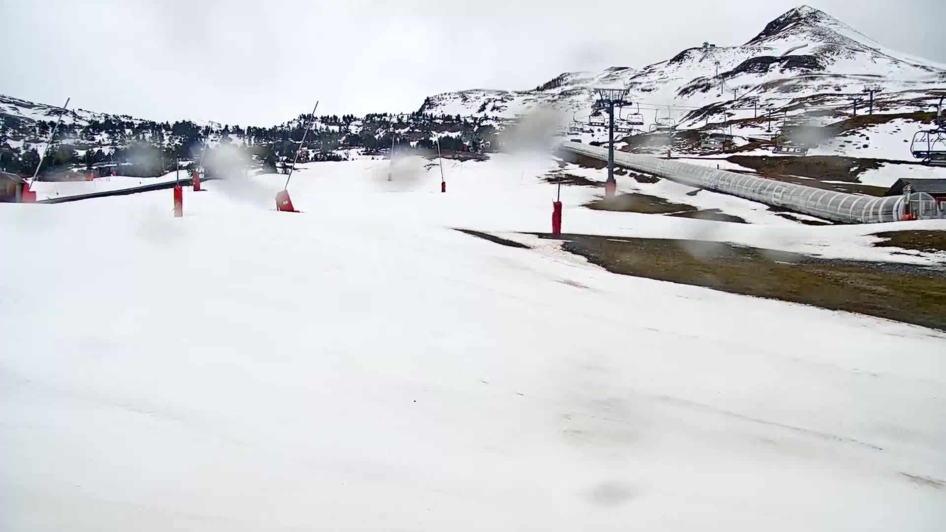 A snow-covered ski slope with a chairlift and snowmaking equipment is visible under an overcast sky with light rain or snow.