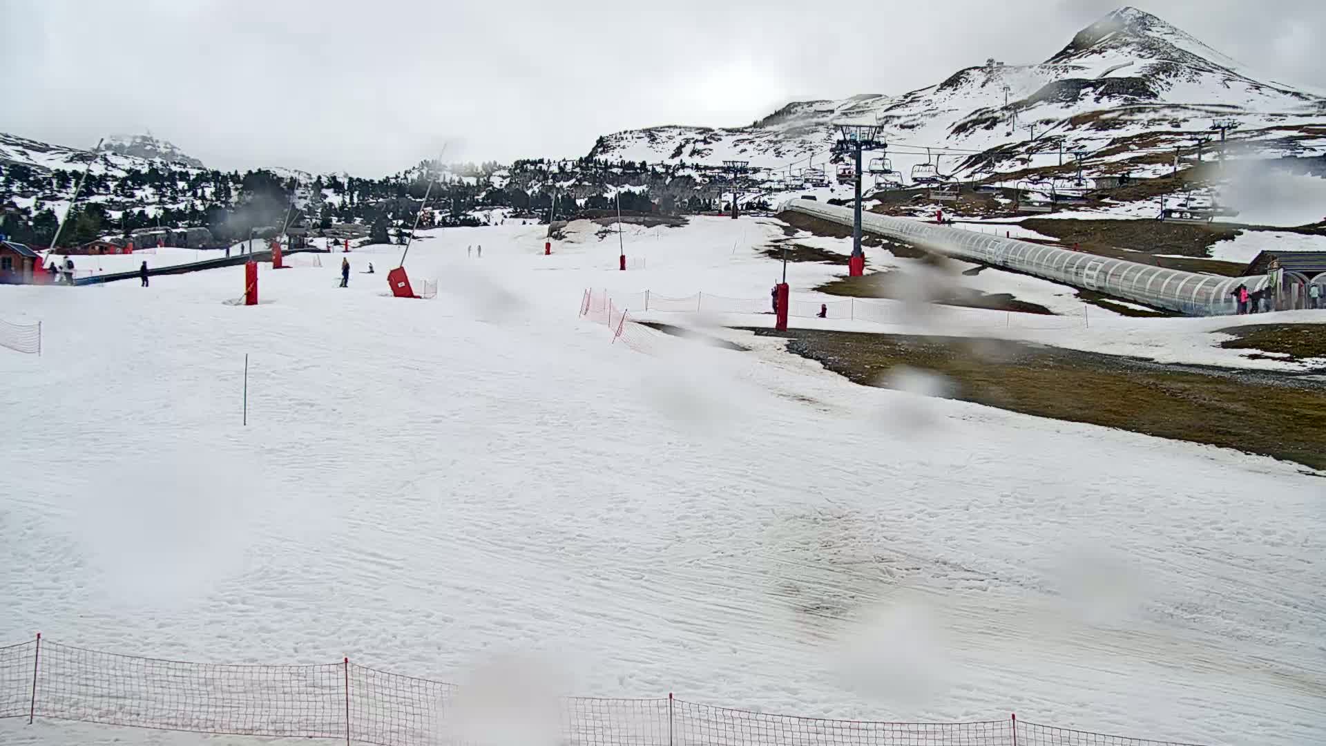 A snow-covered ski slope with a chairlift and a few skiers visible under an overcast sky.