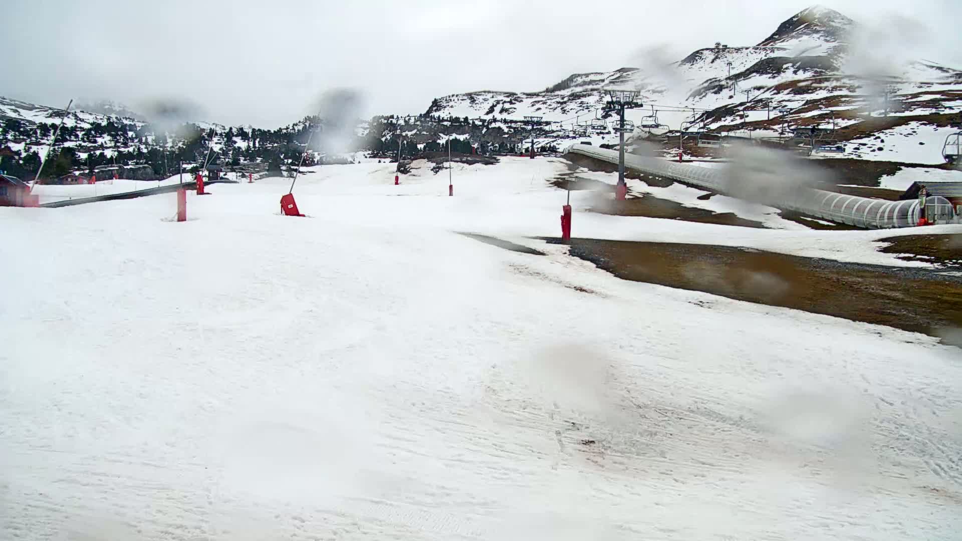 A snow-covered ski slope with a ski lift and several red markers is visible through rain-covered glass, with brown hills and some trees in the background under an overcast sky.