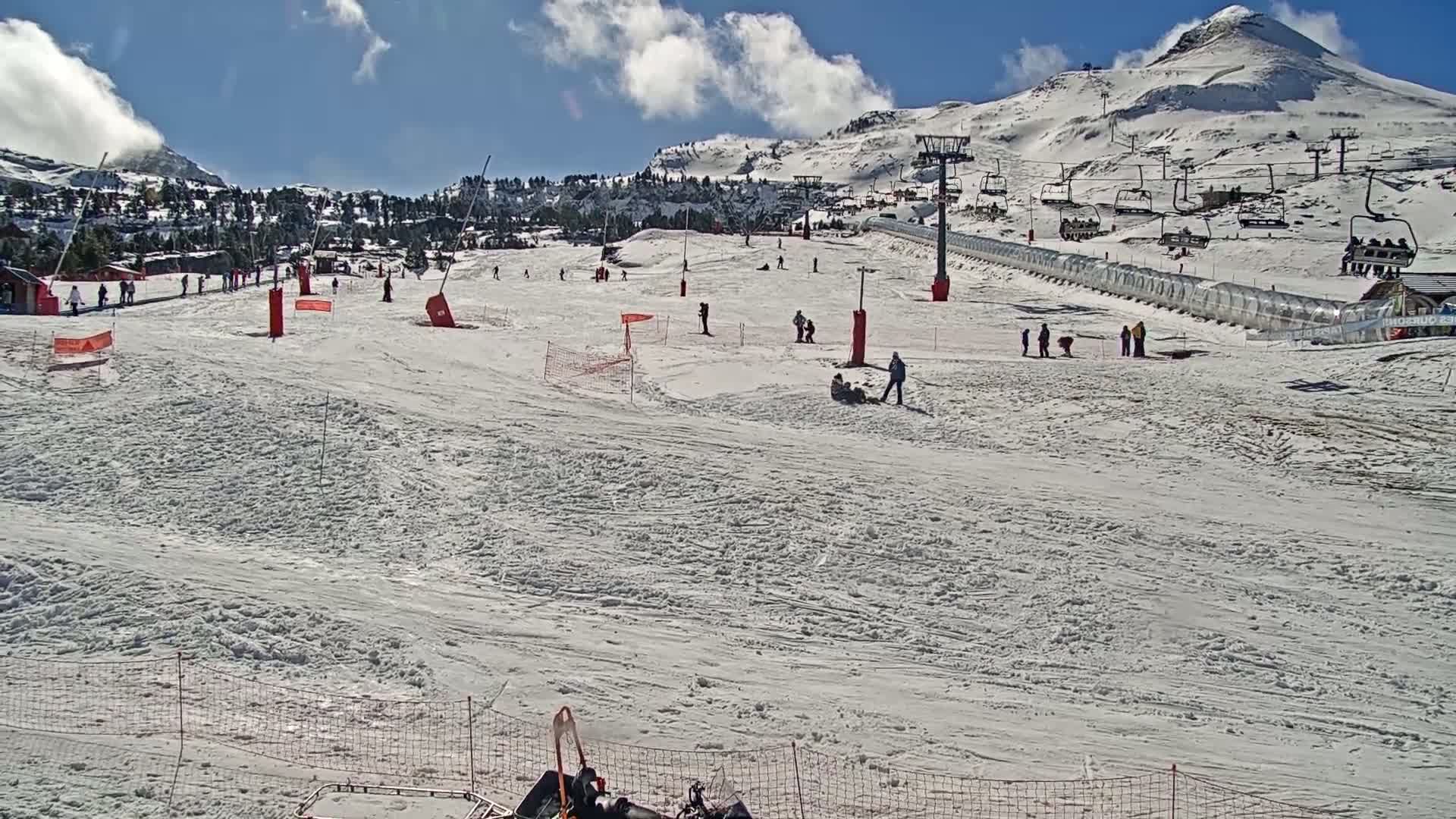 A snow-covered ski slope with several skiers and snowboarders, ski lifts, and a mountain in the background under a mostly sunny sky.