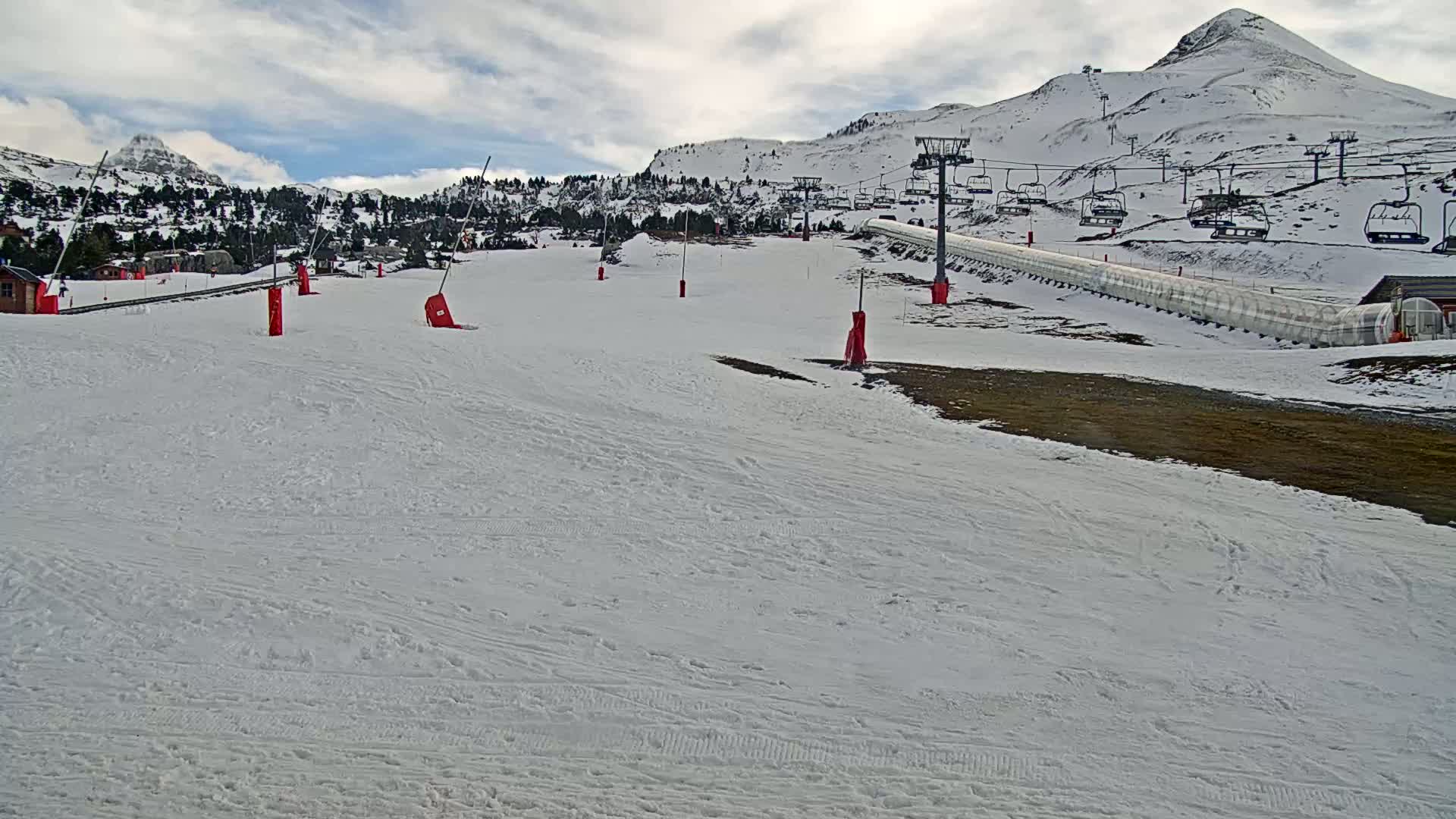 A snow-covered ski slope with several chairlifts and red markers is situated against a backdrop of snow-capped mountains under a cloudy sky.