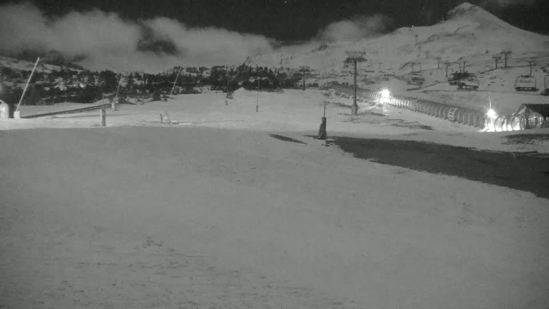 A snow-covered ski resort at night under a cloudy sky shows a row of ski lifts and a building with lights.