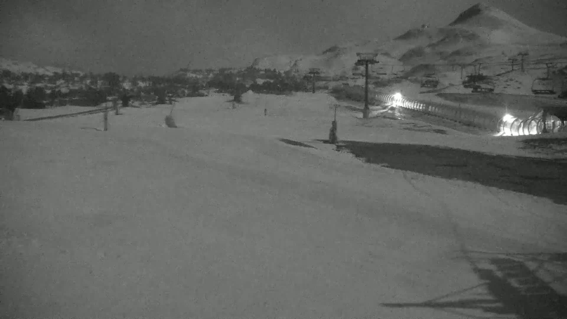 A snow-covered ski slope at night, illuminated by lights near a lift, with snow-covered mountains in the background.