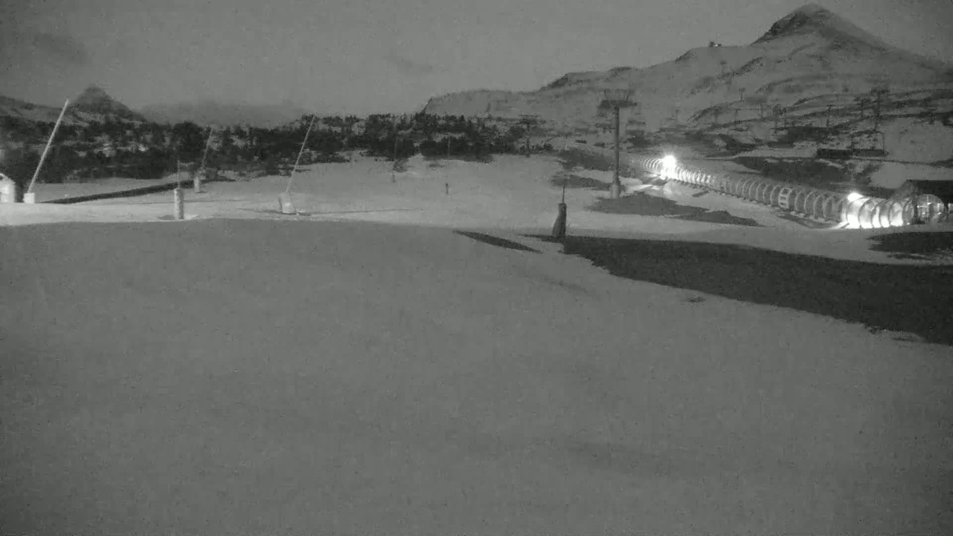 A snow-covered ski slope at night, illuminated by lights near a ski lift and a building in the distance, under a dark, cloudy sky.
