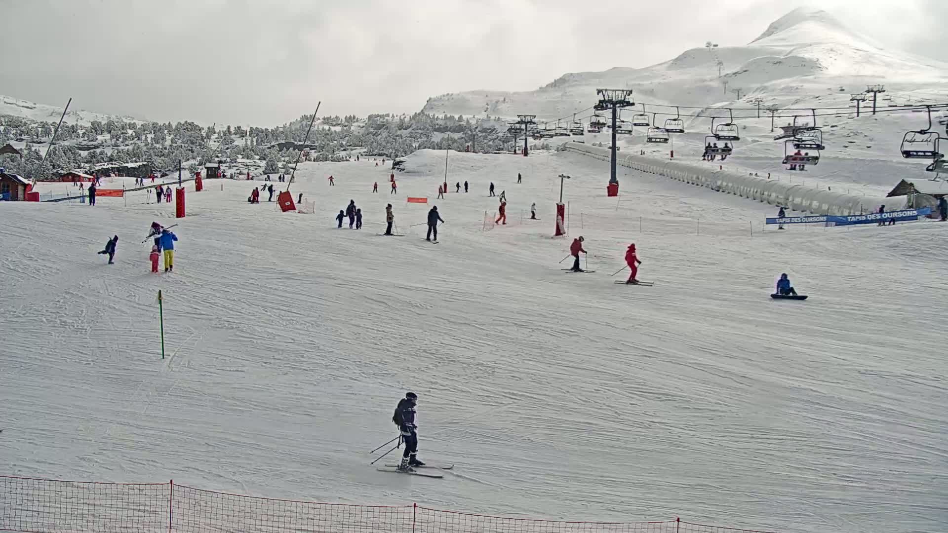A snowy ski slope with several skiers and snowboarders, a chairlift, and a mountain in the background under an overcast sky.