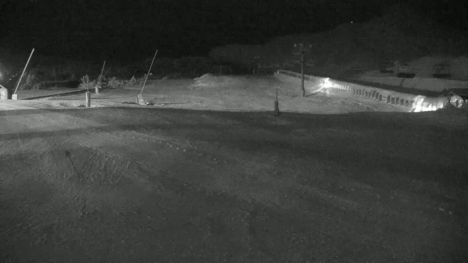 A nighttime, low-light view shows a snow-covered hill and a long, low structure illuminated with lights in the background.