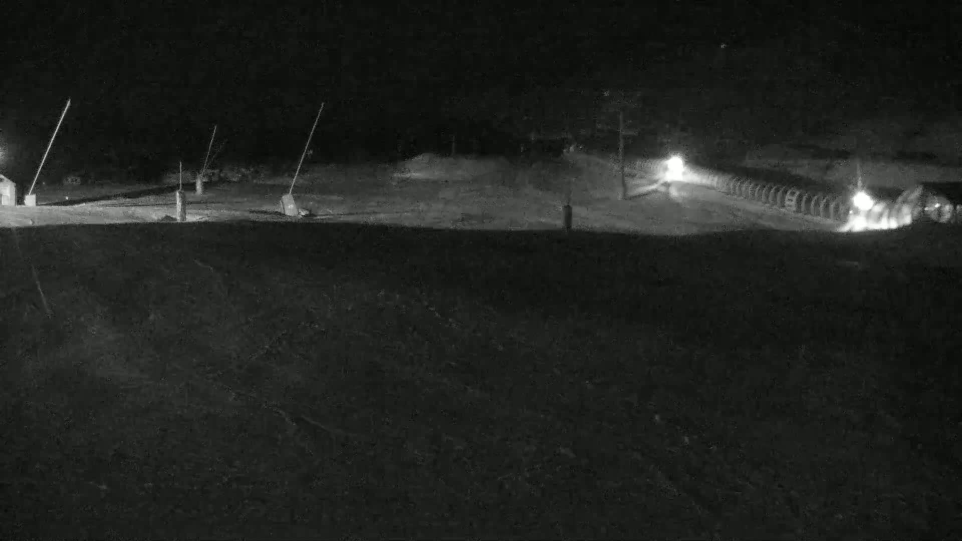 A nighttime, snow-covered scene shows a road with lights and a structure resembling a snow-making machine.