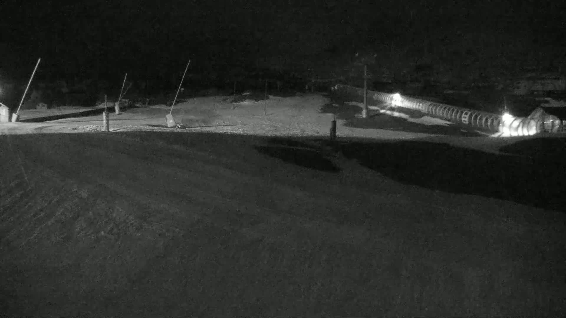 A snow-covered nighttime scene shows a long, illuminated structure, possibly a snowmaking machine, near a sparsely lit area with some poles and small buildings.