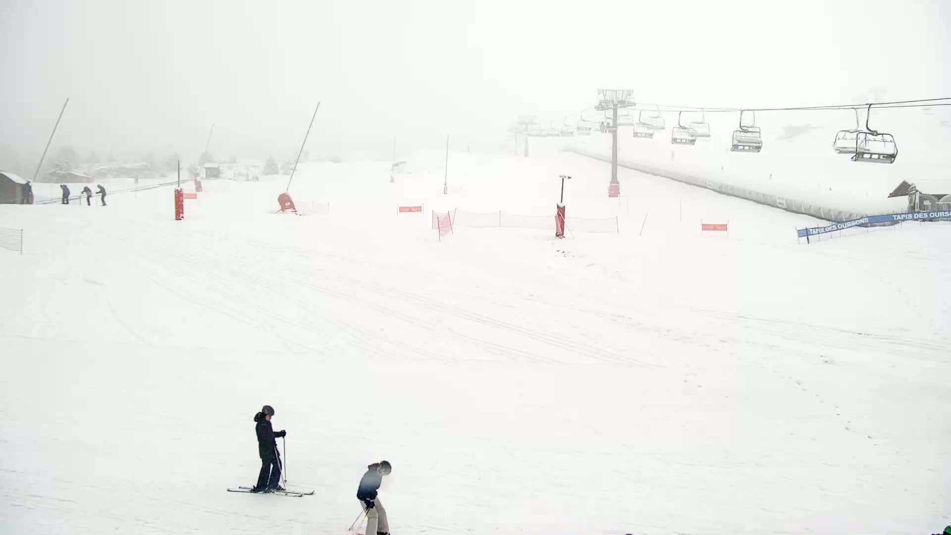 A snowy ski slope with a chairlift and several skiers visible in foggy conditions.