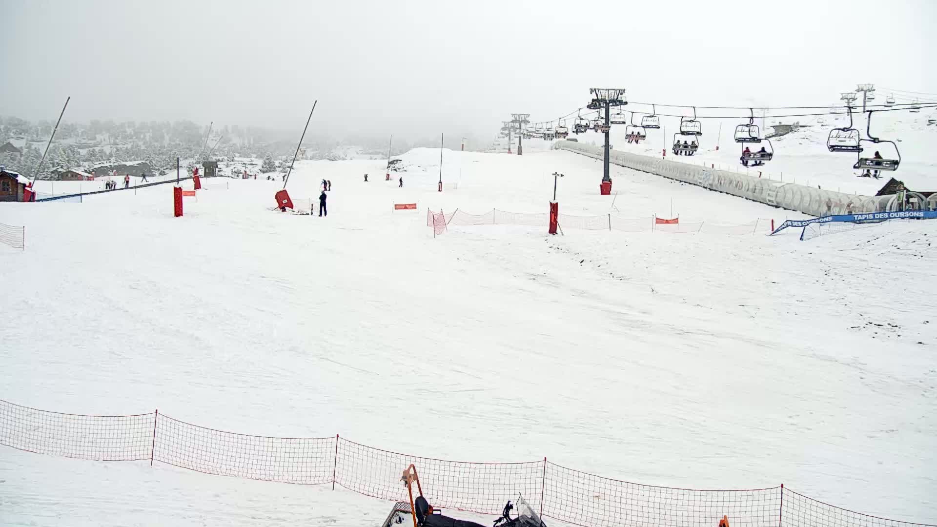 A snow-covered ski slope with a chairlift and a few skiers visible under overcast skies.