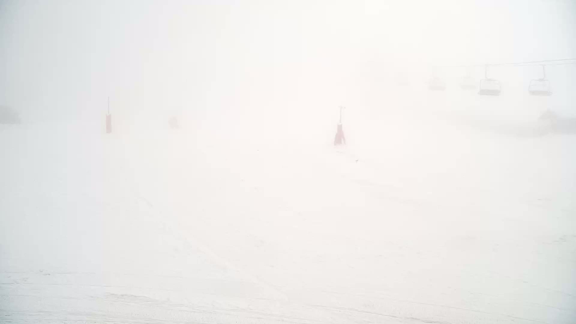A snow-covered ski slope is shrouded in thick fog, with a few ski lifts barely visible in the distance.