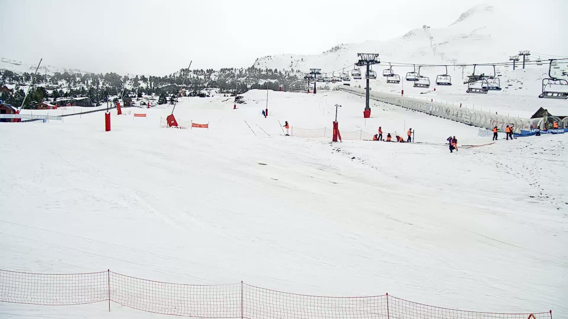 A snowy ski slope with a chairlift and several people working near a snow-covered mountain range under an overcast sky.