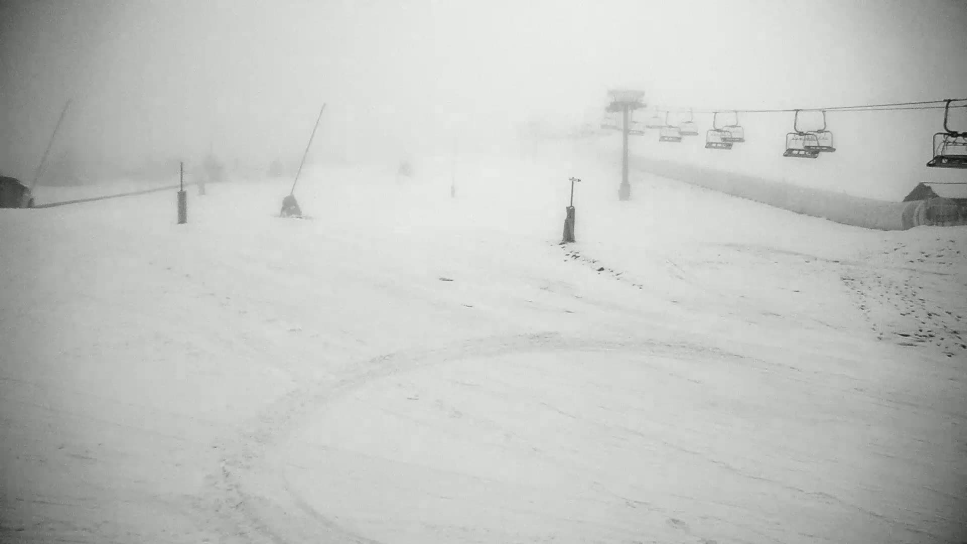 A snow-covered ski slope is shrouded in dense fog, with a chairlift visible in the background.