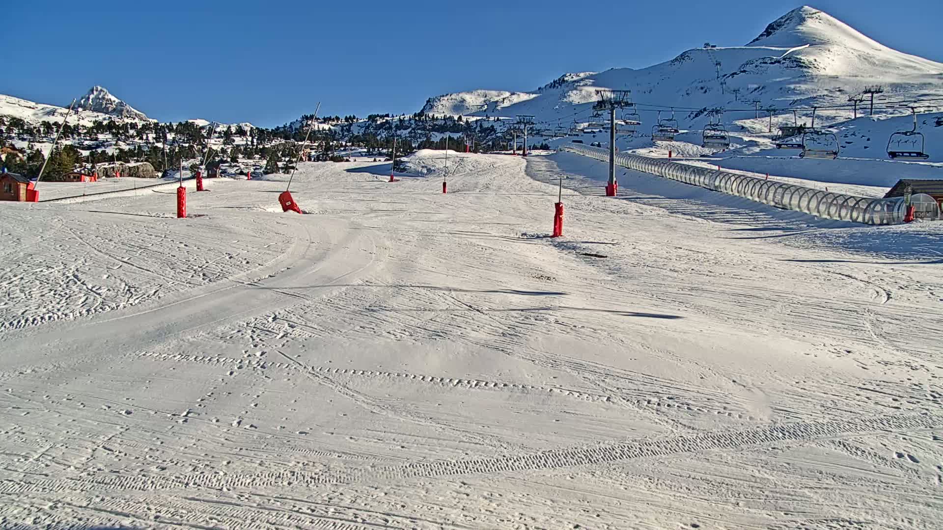 A snow-covered ski slope with a chairlift and several red markers stretches towards snow-capped mountains under a clear blue sky.