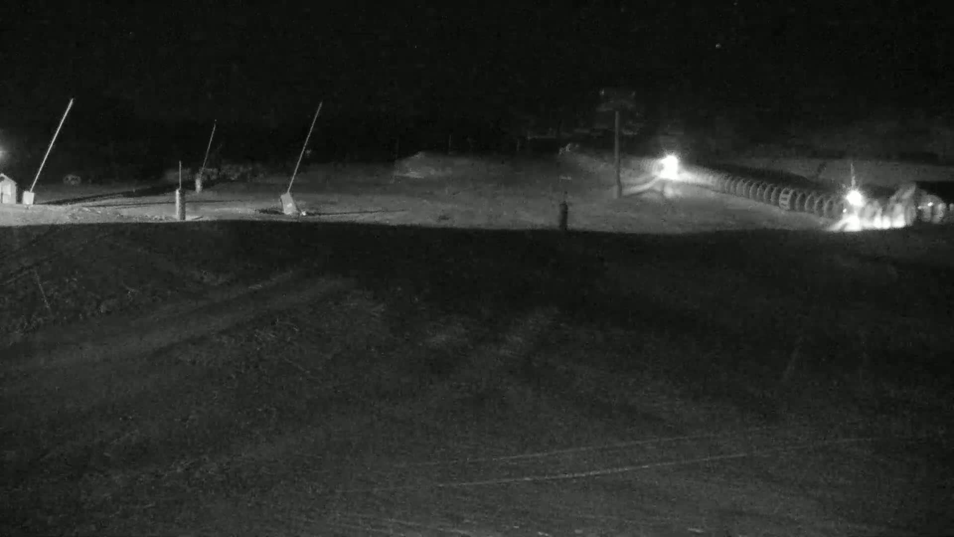 A nighttime, snow-covered scene shows a slope with several illuminated structures and what appears to be a snowmaking system.