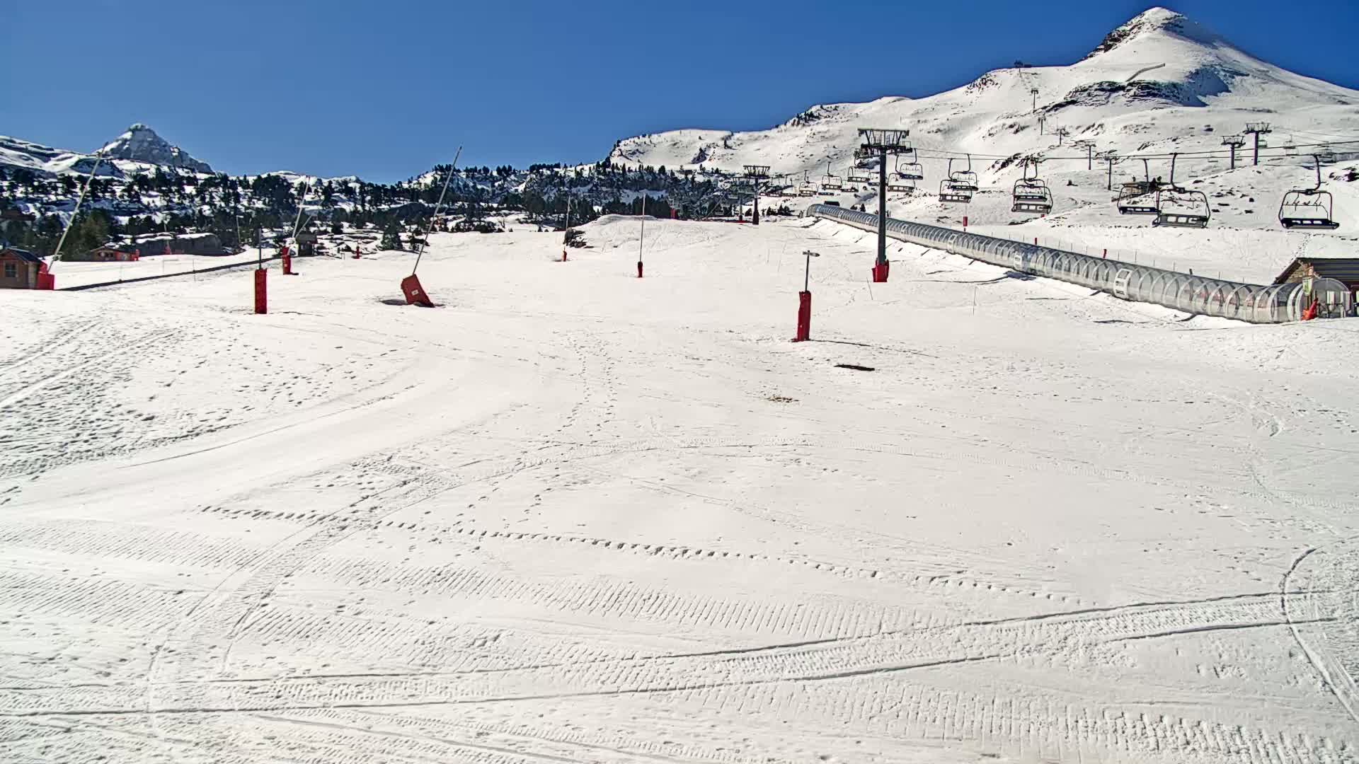 A snow-covered ski slope with a chairlift and a covered conveyor belt extends toward snow-capped mountains under a clear blue sky.