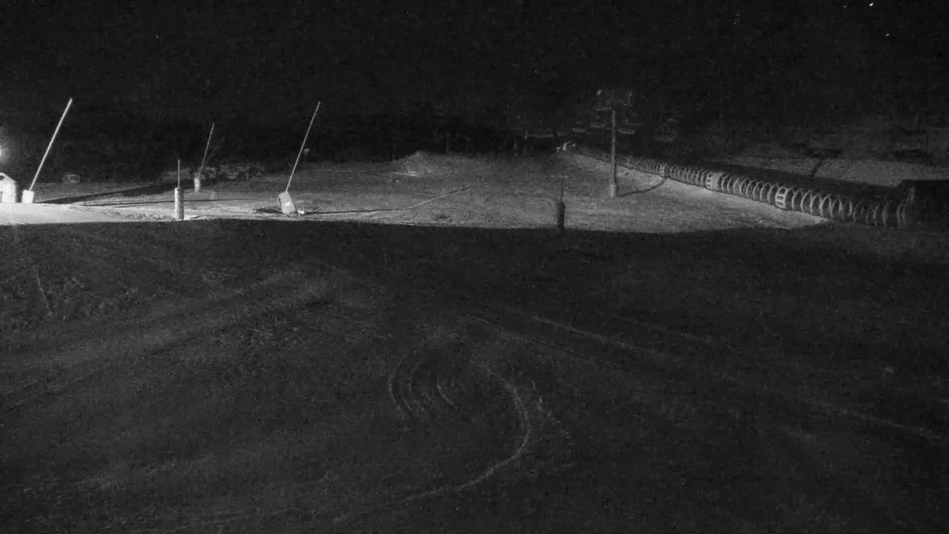 A snow-covered slope at night is illuminated by a single light source, with tire tracks visible on the lower portion of the slope and snowmaking equipment in the background.