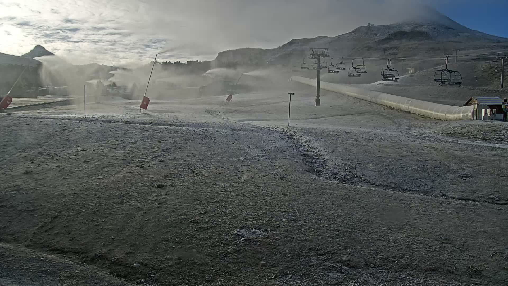 Under a partially cloudy sky, a ski resort prepares for winter with several snow cannons actively spraying artificial snow onto bare ground, while empty chairlifts ascend towards a cloud-shrouded mountain peak.