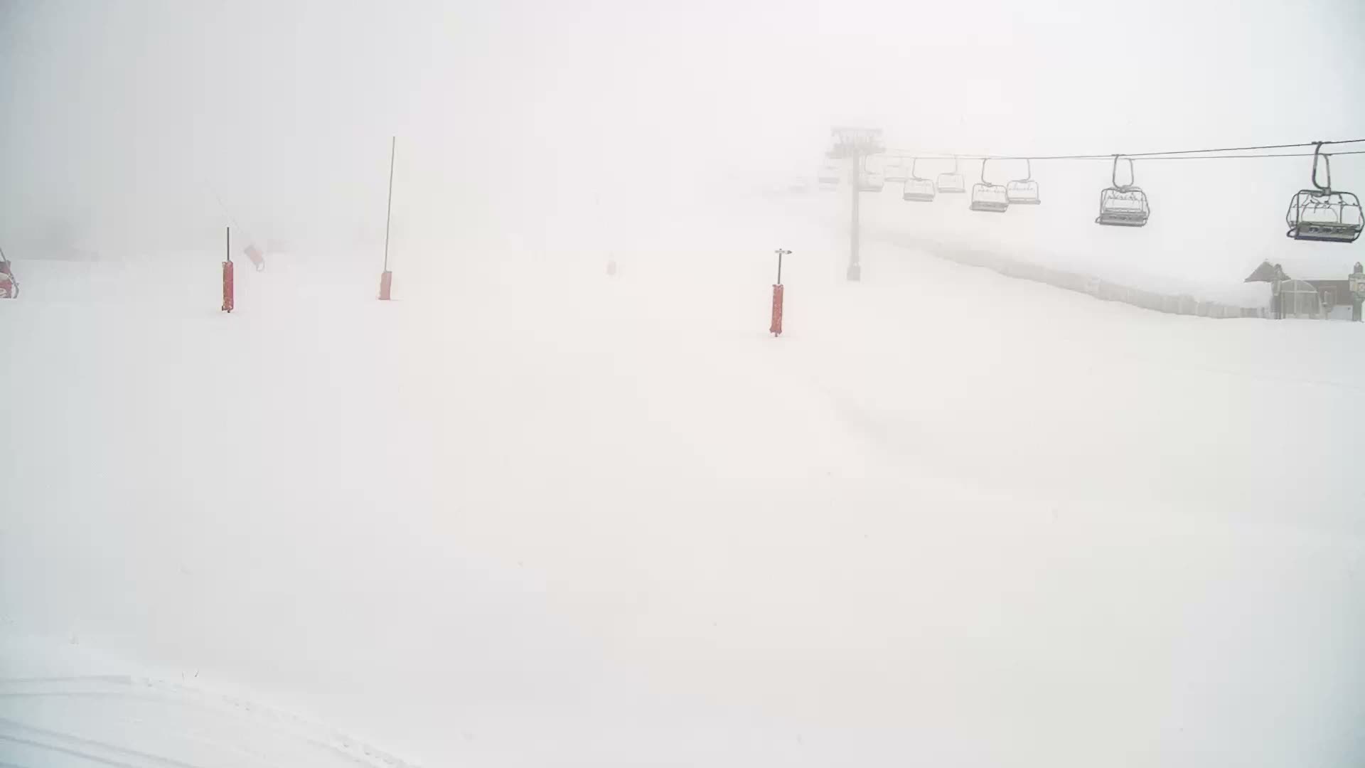 A snow-covered ski slope featuring several red marker poles, a distant ski lift with empty chairs, and a building obscured by dense fog or whiteout conditions.