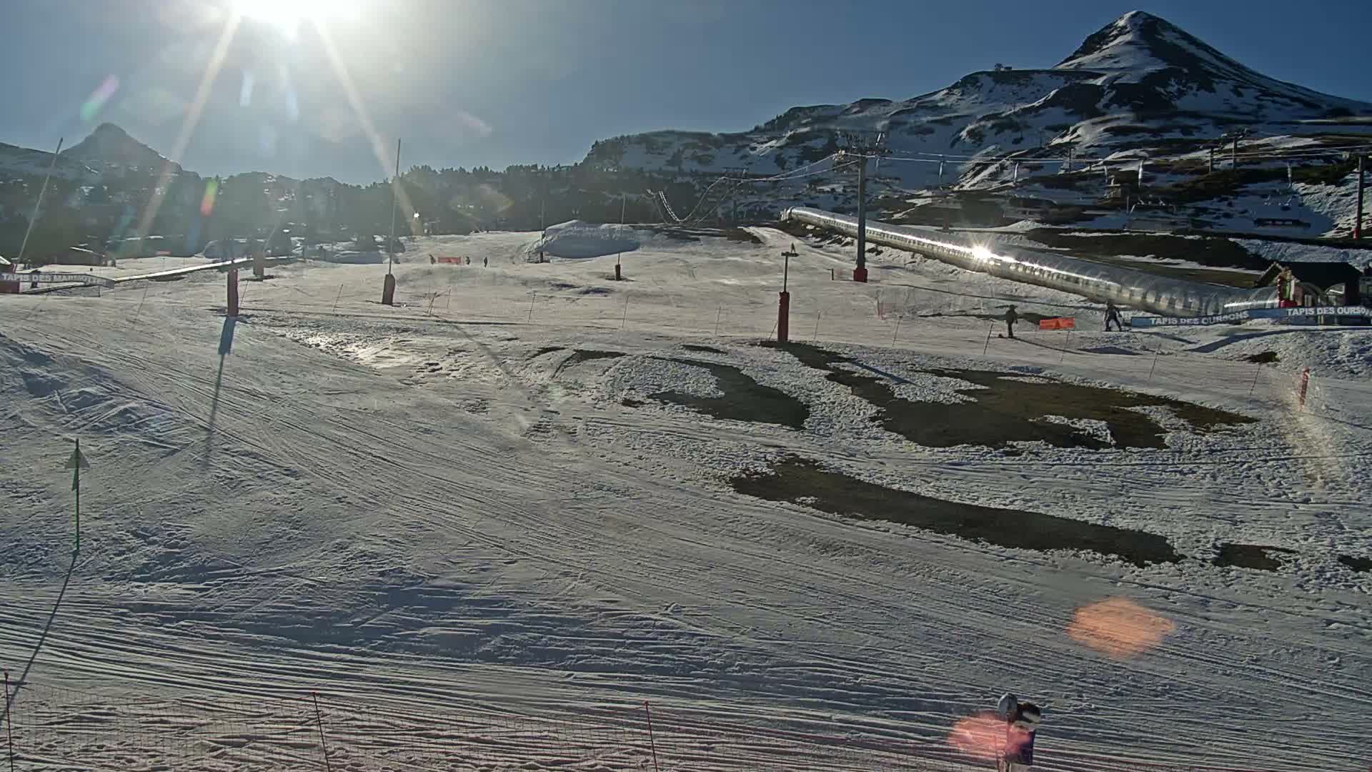 A snow-covered ski slope featuring several red marker poles, a distant ski lift with empty chairs, and a building obscured by dense fog or whiteout conditions.
